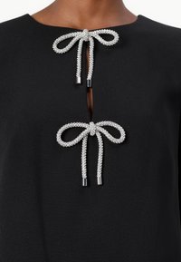 Indumento nero con due dettagli a fiocco in argento con punte metalliche, completato da un'apertura a occhiello centrale. Texture liscia.