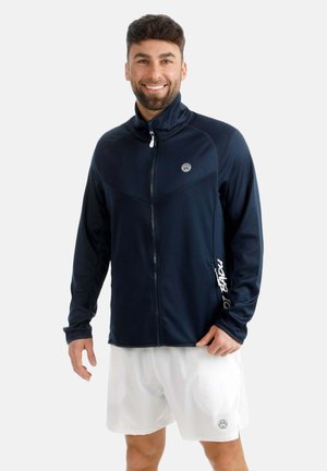 Lächelnder Mann, der eine marineblaue Jacke mit Reißverschluss und weißen Schriftzügen am Ärmel sowie weiße Sportshorts trägt, vor schlichter Hintergrund.