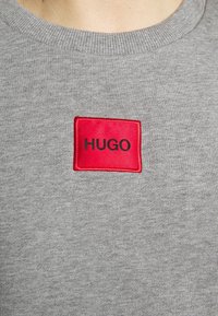 Detailní záběr na šedý svetr s kulatým výstřihem, na kterém se nachází červený obdélníkový nášivka s černým textem "HUGO" umístěným na středu hrudi.