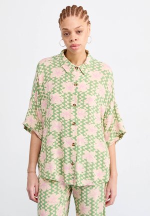 Camicia a maniche corte di colore verde chiaro, con motivi ondulati e fiori rosa. Presenta una chiusura a bottoni sul davanti e un colletto, realizzata in tessuto leggero.