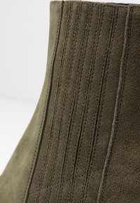 Primo piano di un tessuto in suede verde oliva con cuciture verticali e diagonali che creano un motivo testurizzato.