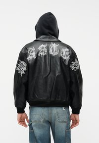 Persona con chaqueta de cuero negra con texto gótico blanco en la espalda y las mangas, capucha negra y pantalones cargo de mezclilla azul.