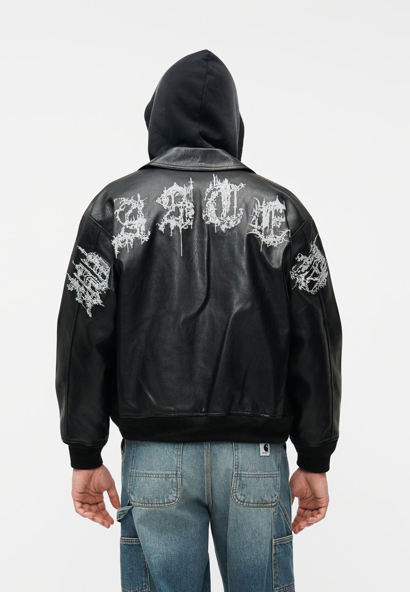 Persona con chaqueta de cuero negra con texto gótico blanco en la espalda y las mangas, capucha negra y pantalones cargo de mezclilla azul.