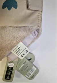 Tejido beige suave con un forro de piel de crema texturizada. Presenta un elemento de diseño en color azul verdoso. Incluye etiquetas de cuidado y de marca en la esquina inferior.