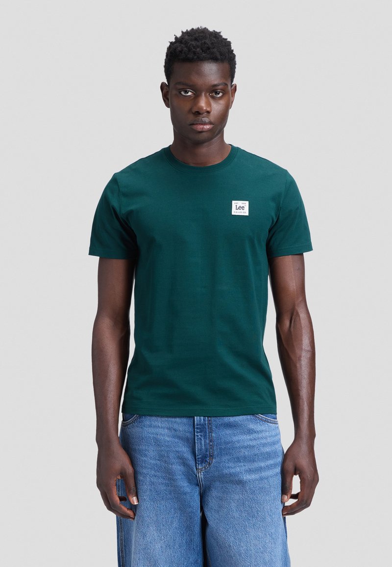 Jeune homme portant un t-shirt vert Lee et un jean bleu en denim, debout devant un fond gris uni.
