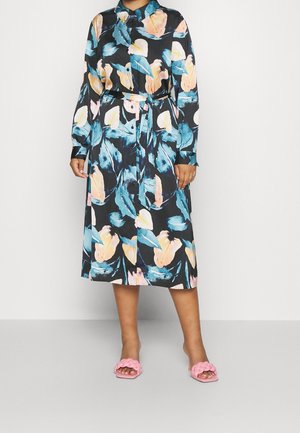 Vrouw draagt een midi-jurk met lange mouwen met een bloemmotief in blauw, roze en perzik en een bijpassende stoffen ceintuur, gecombineerd met roze gevlochten sandalen.