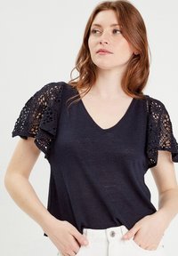 Marineblauw V-hals top van zachte stof, met ingewikkelde kant ruffles aan de mouwen en een ontspannen pasvorm, gecombineerd met lichtgekleurde broeken.