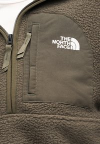 Κοντινό πλάνο σε μπουφάν από φλις ελιάς με τσέπη στο στήθος με φερμουάρ και λευκό κεντημένο λογότυπο "The North Face".