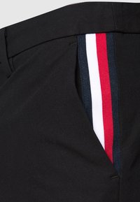 Schwarze Hose mit seitlichem Streifen in Rot, Weiß und Marineblau. Der Stoff wirkt glatt mit einer taillierten Passform und einem Taschendetail.