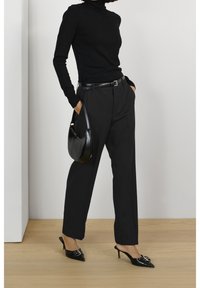 Frau, die einen schwarzen Rollkragenpullover, hochgeschnittene schwarze Hosen, einen schwarzen Gürtel trägt, eine schwarze Handtasche hält und auf Holzfußboden mit spitzen schwarzen Absatzmules steht.