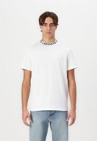 T-shirt blanc à manches courtes avec un col noir côtelé, arborant le texte "LACOSTE". Tissu en coton doux, coupe ajustée, logo minimaliste sur la poitrine.