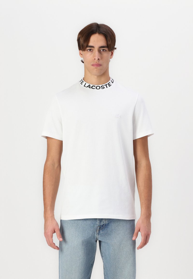 T-shirt blanc à manches courtes avec un col noir côtelé, arborant le texte "LACOSTE". Tissu en coton doux, coupe ajustée, logo minimaliste sur la poitrine.
