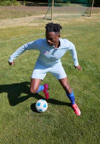 Light blue athletic soccer kit with pink accents, featuring a long-sleeve top and shorts. Player dribbling a soccer ball on green grass.