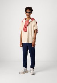 Tricou din bumbac bej, pantaloni sport bleumarin și adidași gri. Hanorac roșu aruncat pe umeri. Modelul poartă ochelari de soare negri.