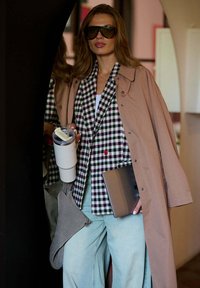 Žena v nadměrných slunečních brýlích a béžovém trenčkotě, která drží bílý tumbler a laptop, stojí uvnitř poblíž zrcadla.