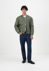 Olijfgroene bomberjack met geribbelde manchetten, ritssluiting aan de voorkant en een patch op de linkermouw. Gecombineerd met donkerblauwe denim jeans en zwarte schoenen.