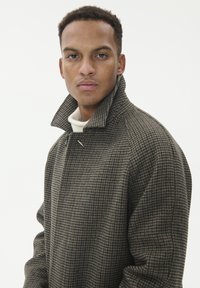 Cappotto a motivo houndstooth marrone con colletto alto, caratterizzato da una leggera lucentezza e una vestibilità morbida e rilassata. Indossato sopra un maglione bianco.