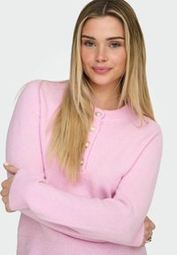 Pull en maille rose clair avec un col arrondi, doté d'une patte de boutonnage avec des boutons nacrés et des poignets côtelés. Texture douce.