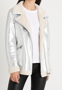 Giacca da motociclista in argento metallizzato con collare e polsini in pelliccia sintetica color crema, dotata di tasche con zip e design accorciato.