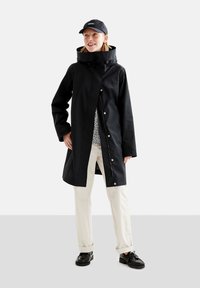 Veste imperméable noire avec un col haut et des fermetures à pression, associée à un pantalon beige clair et des chaussures noires. Le modèle porte une casquette noire.