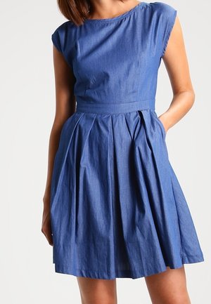Mujer con vestido vaquero azul sin mangas con corpiño ajustado y falda plisada en línea A, una mano en el bolsillo, sobre un fondo claro y liso.