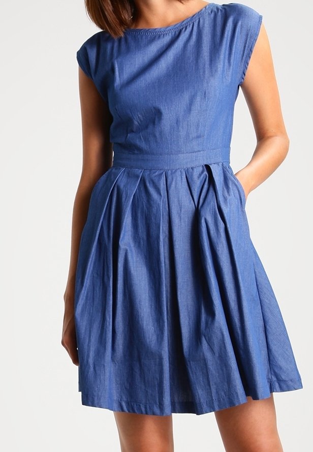Femme portant une robe en denim bleu sans manches avec un corsage ajusté et une jupe plissée en ligne A, une main dans la poche, sur un fond clair uni.