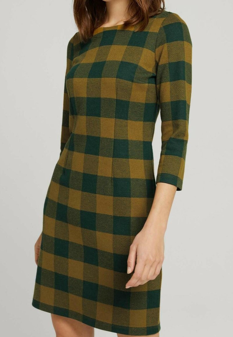 Femme portant une robe à carreaux verts et bruns, longueur genoux, manches trois-quarts, debout devant un fond clair uni.