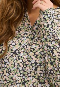Camicia a bottone floreale con sfondo scuro, caratterizzata da motivi rosa, blu e verdi. Maniche lunghe con polsini a bottone, tessuto leggero.