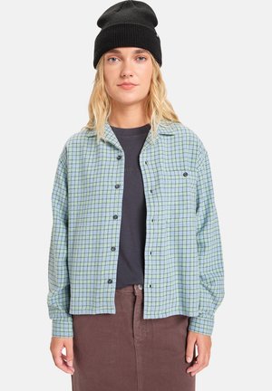Geruit overhemd in lichtblauw en groen, met een losse pasvorm en een enkele borstzak, gecombineerd met een donker T-shirt en een bruine rok.