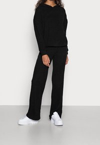 Schwarzer Kapuzenpullover aus Fleece und passende Hose, weiche Textur, lockere Passform, getragen mit klobigen weißen Sneakers auf einem hellen Boden.