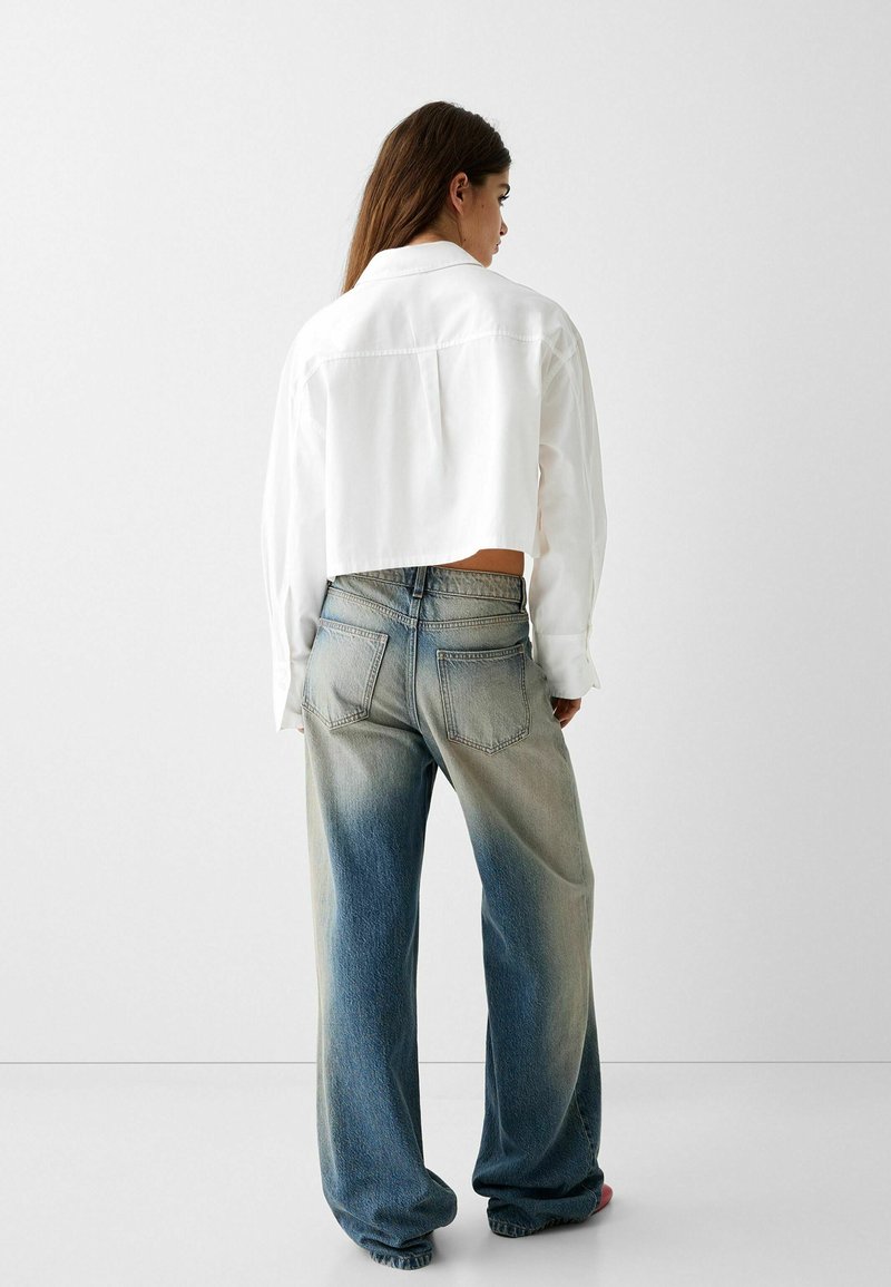 Camicia bianca cropped abbinata a jeans a gamba larga blu sbiaditi, con una transizione graduale di colore dal chiaro allo scuro nella parte inferiore.