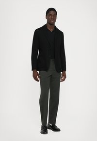 Mand iført sort blazer, sort polo-shirt, mørkegrå bukser og sorte habitstøvler, står foran en ensfarvet lys baggrund.