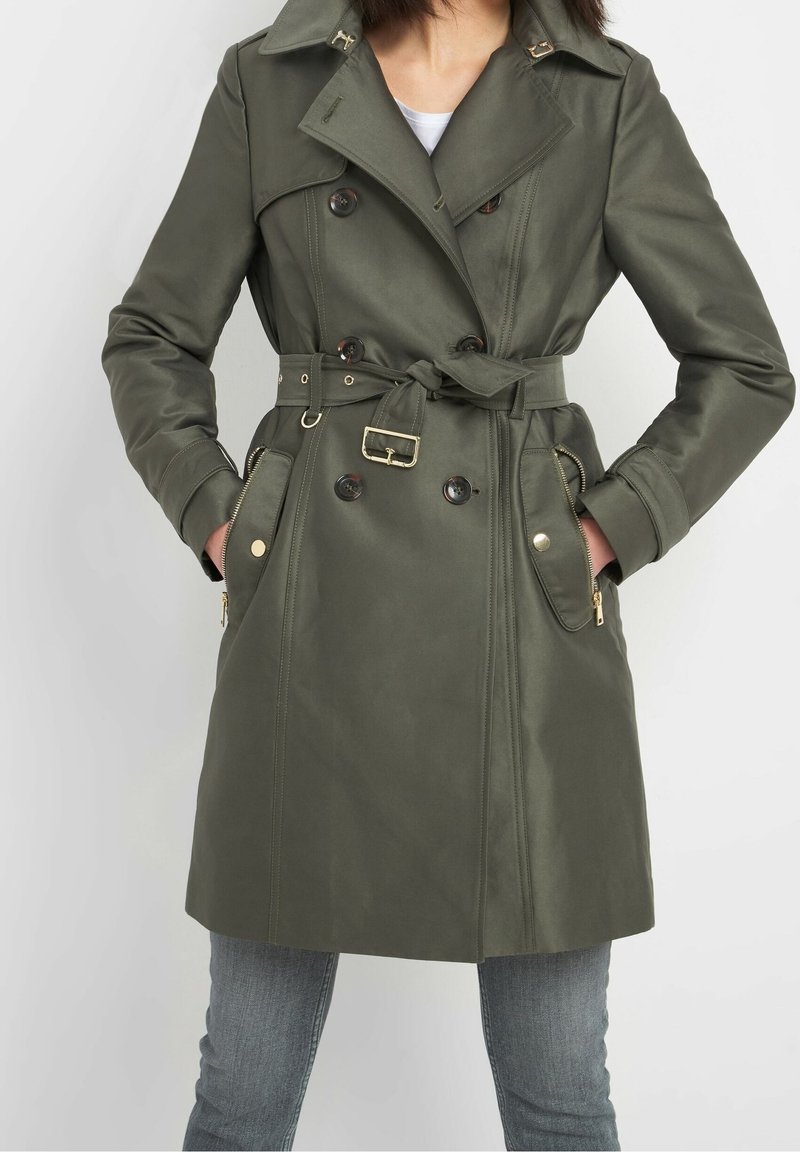 Femme portant un trench-coat mi-long olive à double boutonnage avec ceinture, poches à rabat boutonnées et poches latérales zippées, mains dans les poches.