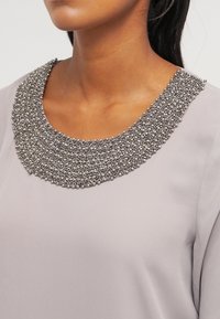 Grijze top met een textuur, bewerkte kraag met metalen accenten en een gladde stofafwerking, met een ronde halslijn.