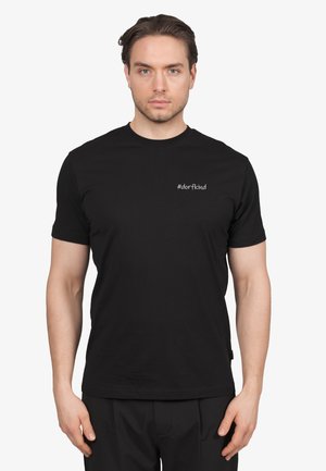 Hombre con cabello corto castaño que lleva una camiseta negra con el texto "#dorfkind" en blanco en el lado izquierdo del pecho, de pie frente a un fondo blanco.