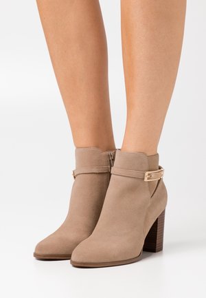 Béžové kotníkové boty z broušené kůže s špičatou špičkou, zlatými kovovými doplňky a naskládaným dřevěným podpatkem. Mají boční zip pro snadné oblékání.
