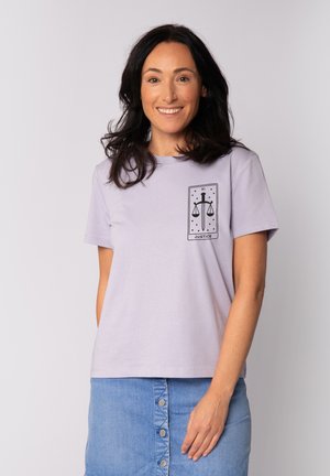 T-shirt en coton lavande à manches courtes, présentant un graphique noir représentant les échelles de la justice avec le mot "JUSTICE" en dessous sur le côté gauche.