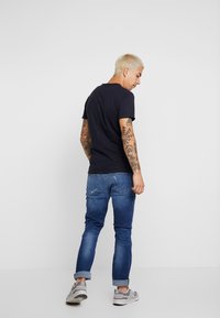 Muž s krátkými blond vlasy a rukávovými tetováními, oblečený v černém tričku, modrých džínech s ohrnutými nohavicemi a šedých teniskách, stojící na bílém pozadí.