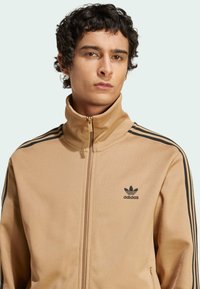 Bež jakna na patent s glatkom tkaninom, koja ima crne tri pruge na ramenima i crni Adidas logo na prsima.