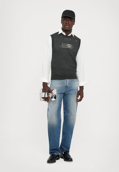 Mand iført sort kasket, hvid skjorte, mørkegrå ærmeløs sweater med kalenderprint, bleget blå jeans, sorte sko, som holder en reflekterende sølv clutch.