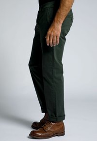 Pantalones de algodón verdes con un corte ajustado, puños enrollados y un bolsillo frontal. Llevados con botas de cuero marrón y una camisa negra.
