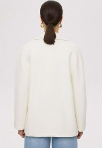 Witte wolmix oversized jas met een klassieke kraag en rechte snit. Voorzien van lange mouwen en zichtbare naden; gedragen over lichtblauwe jeans.