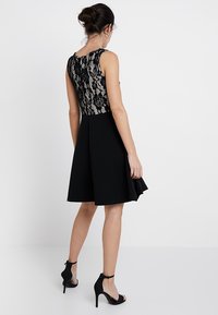 Robe noire avec un corsage en dentelle ajusté et une jupe évasée. Présente une texture lisse et un ourlet au genou. Sandales à talons assorties.