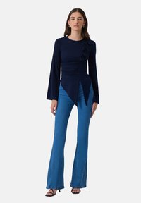Navy long-sleeve top with side ruching and decorative bows, paired with flared blue jeans and black strappy heels. Smooth fabric texture.