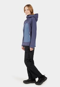 Violette und blaue Zip-Jacke mit Kapuze, ausgestattet mit Reißverschlusstaschen und ergonomischer Passform, kombiniert mit schwarzen Cargo-Hosen und Schuhwerk.