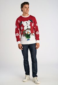 Czerwona świąteczna sweter z białymi płatkami śniegu i wzorem bałwanka z zielonym pomponem. Zestawiony z ciemnymi dżinsami i białymi trampkami.