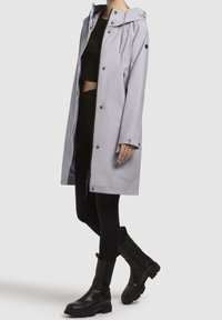 Femme portant un imperméable à capuche gris clair par-dessus un crop top noir, des leggings et des bottines noires épaisses, debout avec une jambe croisée derrière.