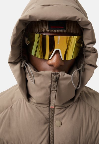 Beige insulated jacket with a high collar, zipper, and drawstrings. Gold mirrored sunglasses and a brown knit beanie complete the look.