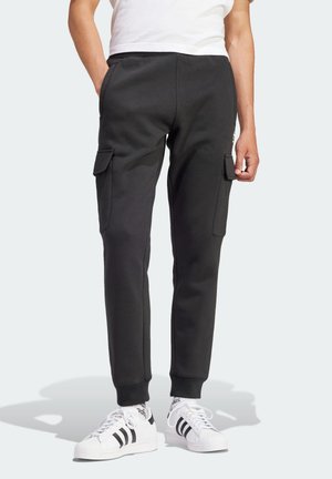 Personne portant un pantalon cargo jogger noir avec des poches à rabat et des baskets Adidas blanches avec des rayures noires, debout sur un fond clair.