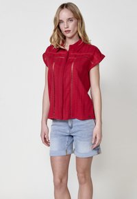 Rode shirt met korte mouwen en een kraag, voorzien van uitsnijdingen aan de voorkant, gecombineerd met lichtblauwe opgerolde denim shorts.